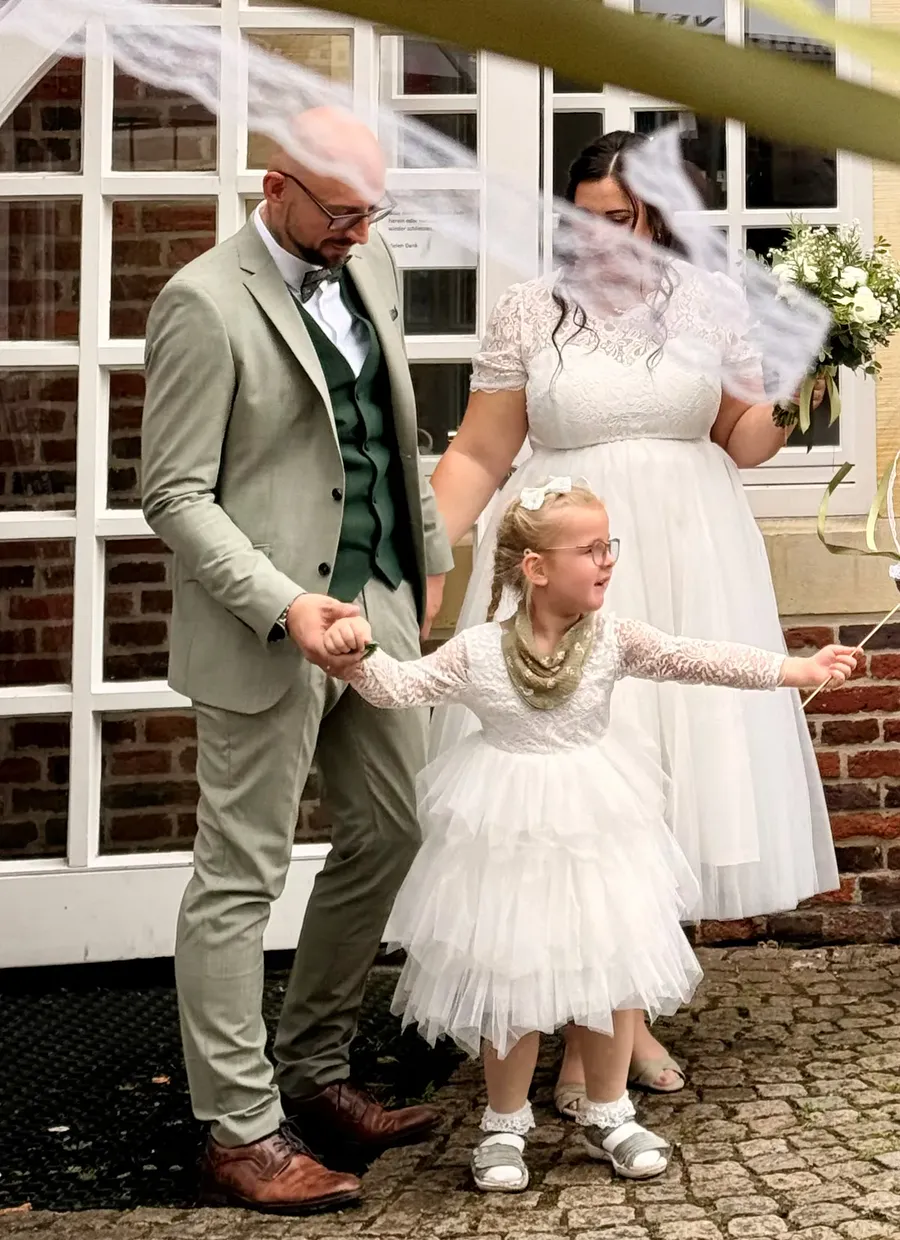 Unsere eigene Hochzeit: Das Brautpaar tritt mit einem kleinen Mädchen an der Hand aus dem Standesamt, über ihnen fliegen Luftschlangen